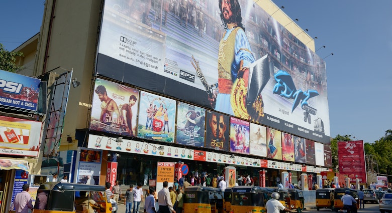 Yellow motorized rickshaws and pedestrians move down the street in front of a movie theater in Chennai plastered with movie posters, including a large multi-story ad featuring a male movie star with long hair and traditional Indian costume.
