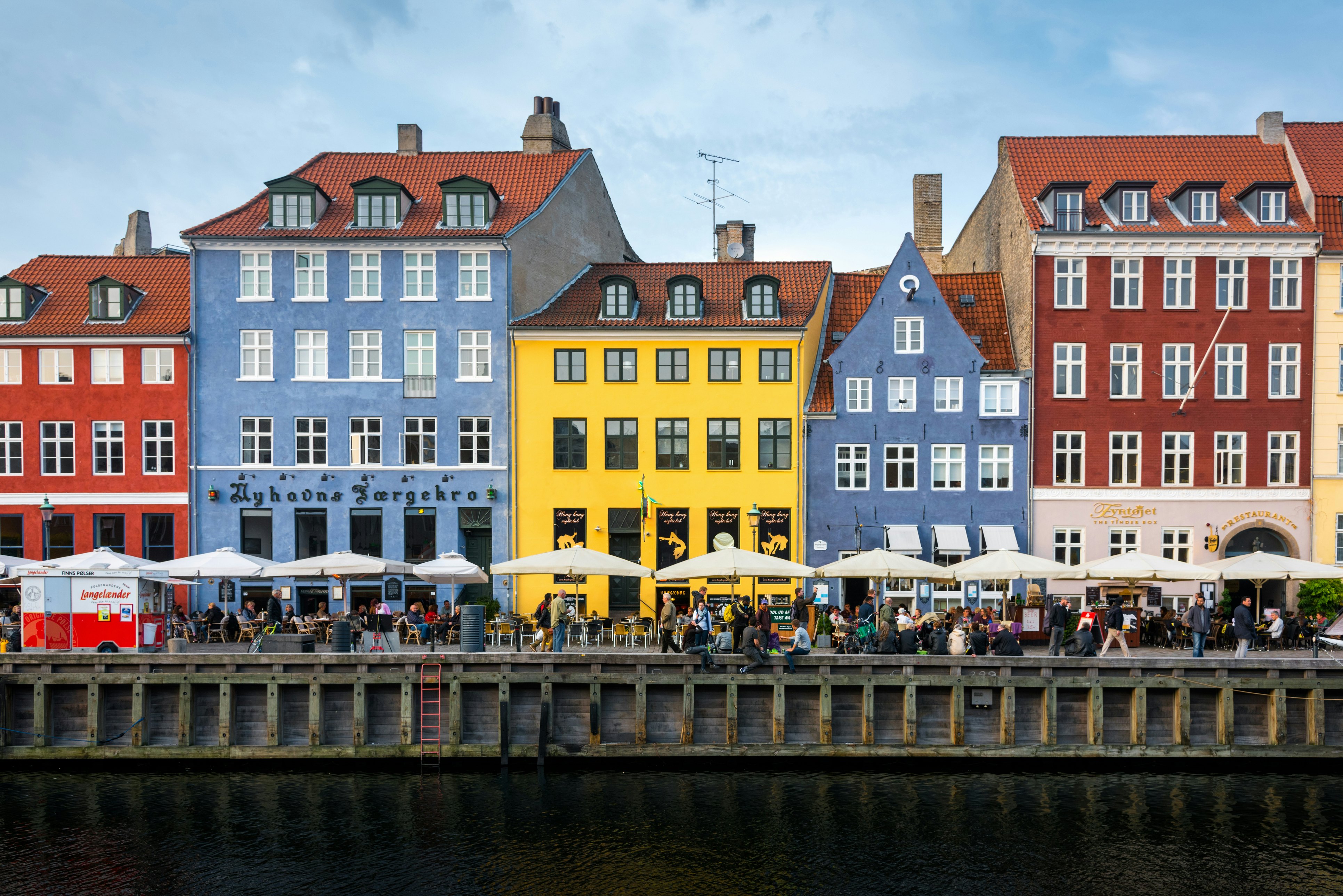 Red, blue and yellow buildings line a waterway; people are sitting at cafes under umbrellas along the waterfront.
