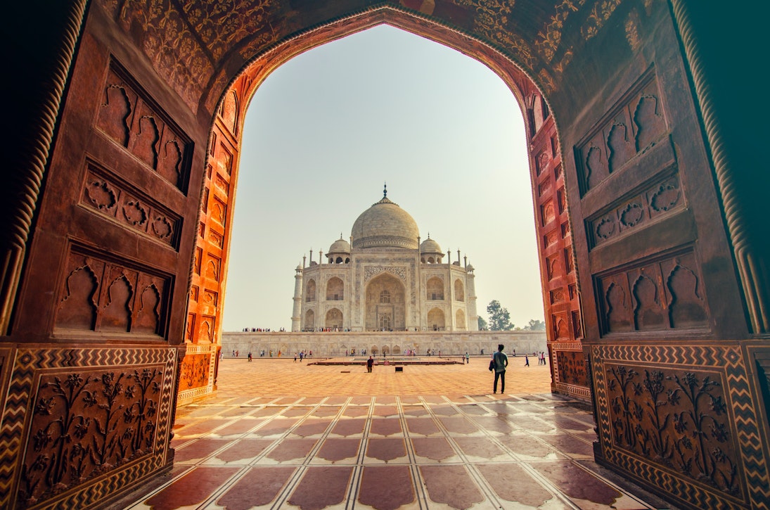 The Taj Mahal as viewed through the doorway of the main gate, which frames the building neatly. Crowds walk towards the white marble structure, which has a domed roof and four pillars surrounding it.