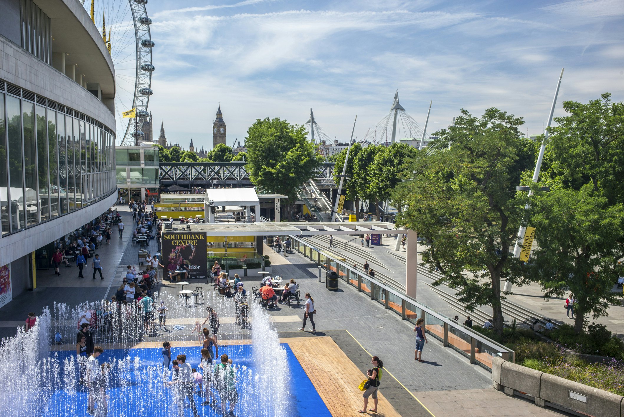 A terrace above a walkway on a sunny day with kids splashing in water fountains