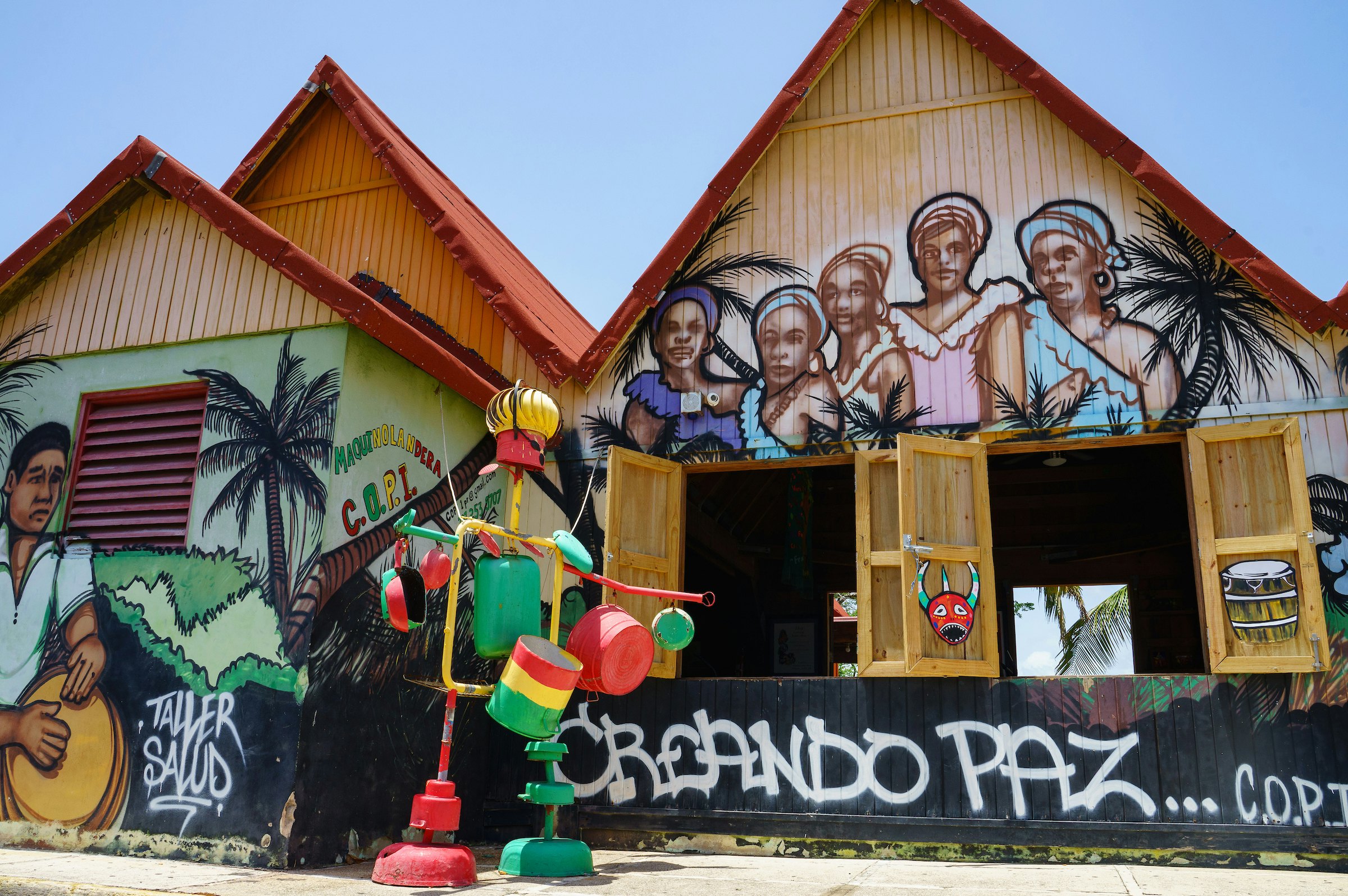 A pair of buildings are covered with images of women with head scarves and a man playing a drum; in front of the building is a colorful structure made of pots and pans; Sustainable Puerto Rico