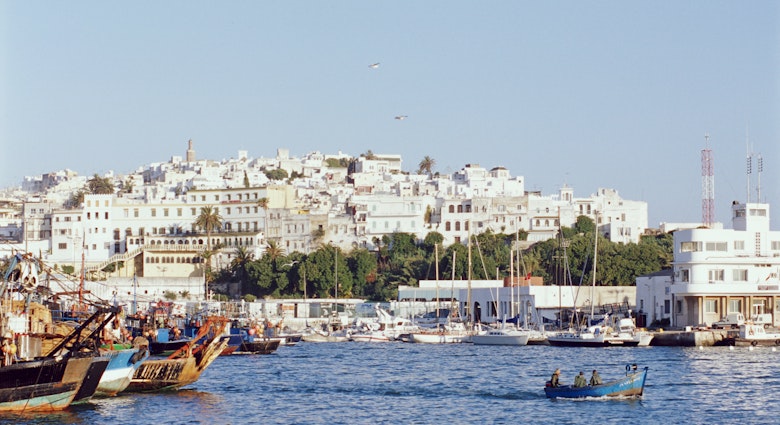 Looking over the waters of the Mediterranean, the white facades of Tangier's old city rises from the shoreline; boats are moored to shore, while a small one makes its way across the harbour.