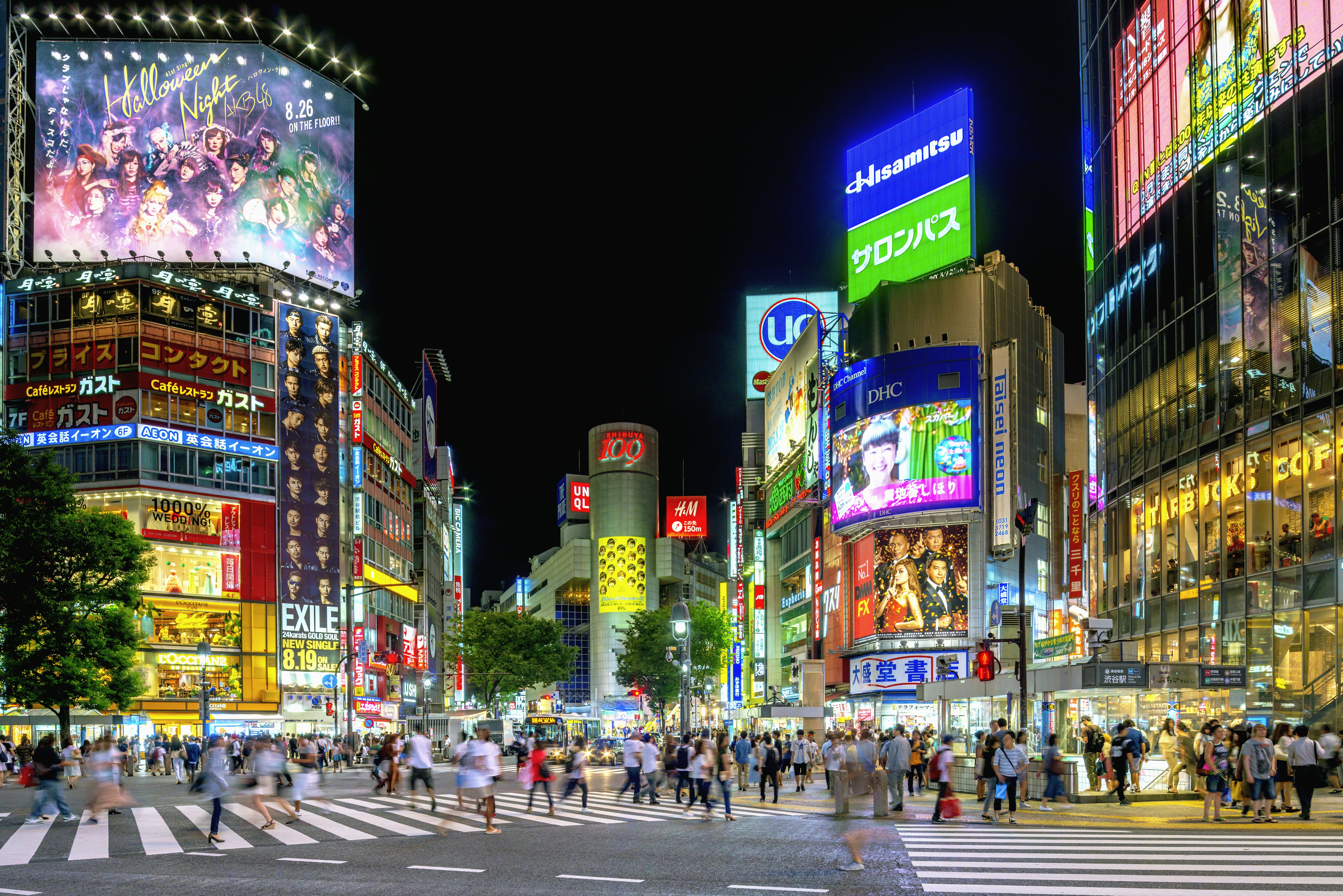 People crossing a large crossroads at night with neon signs lit up on the buildings all around.