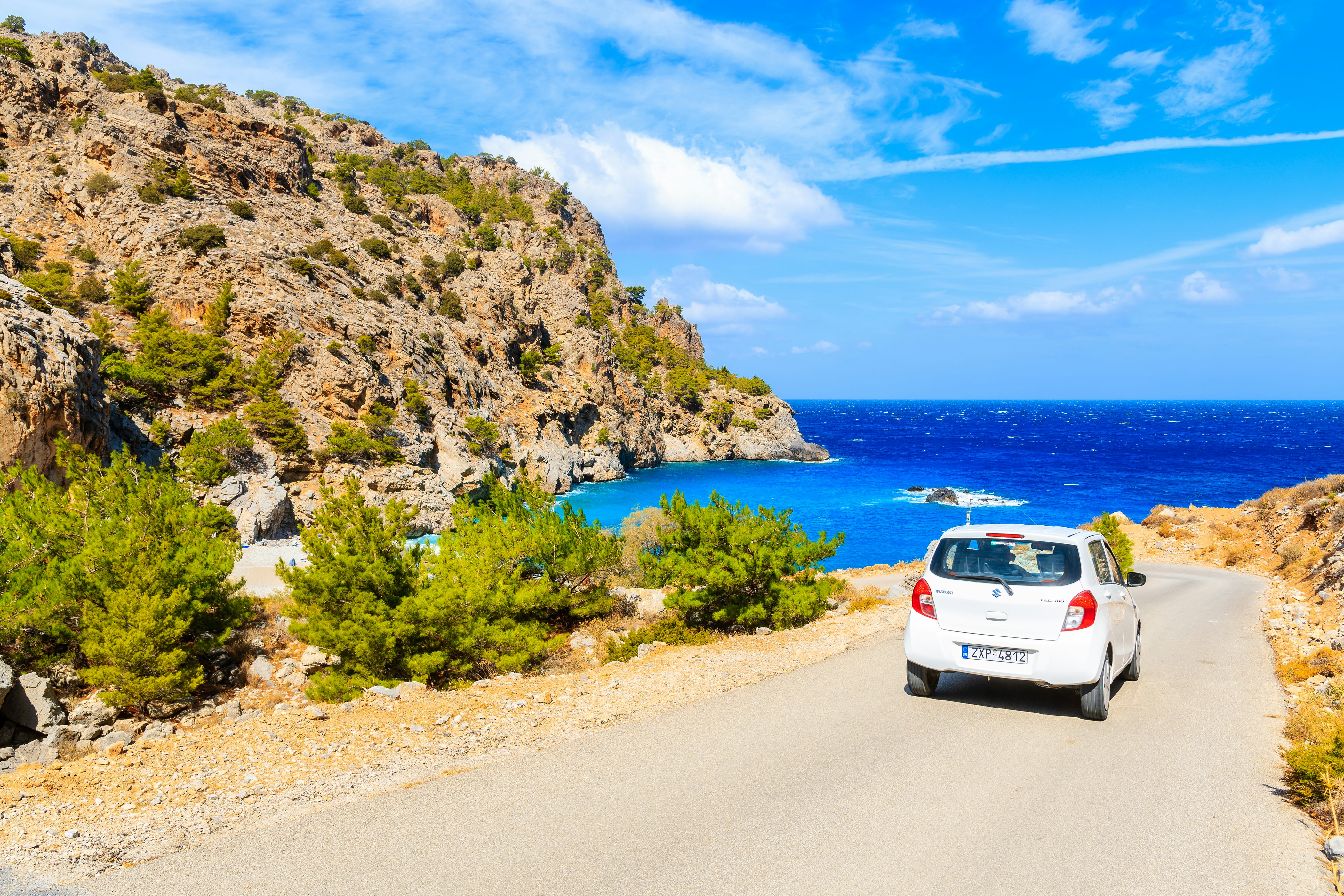 A small white car on a narrow paved road near a cove. A rocky slope rises to the left, while deep blue water is seen at sea.