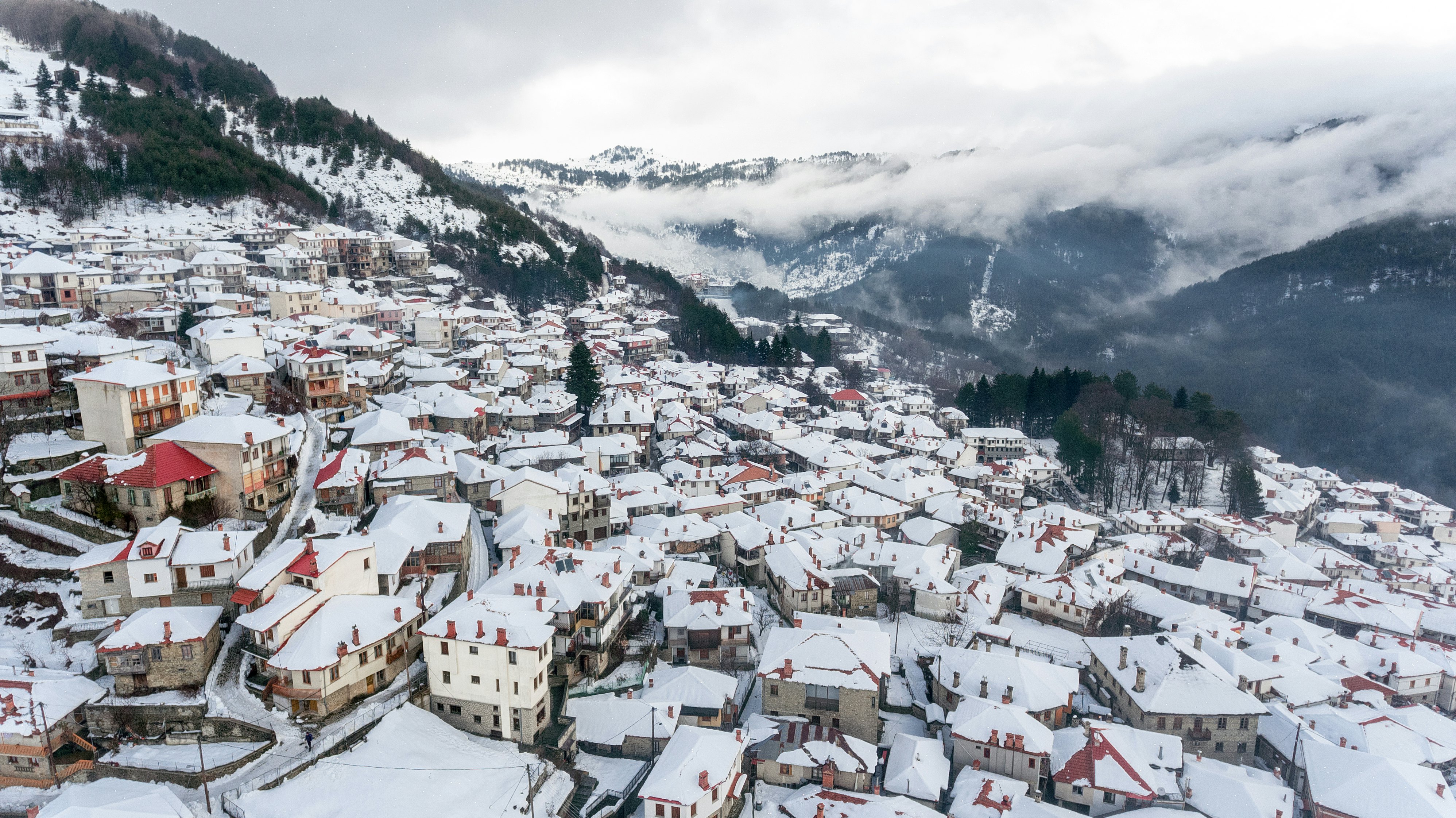An aerial view of a mountain village after a snowstorm. Fresh white snow covers the roofs of the town, and misty clouds hug the mountain slopes in the distance.