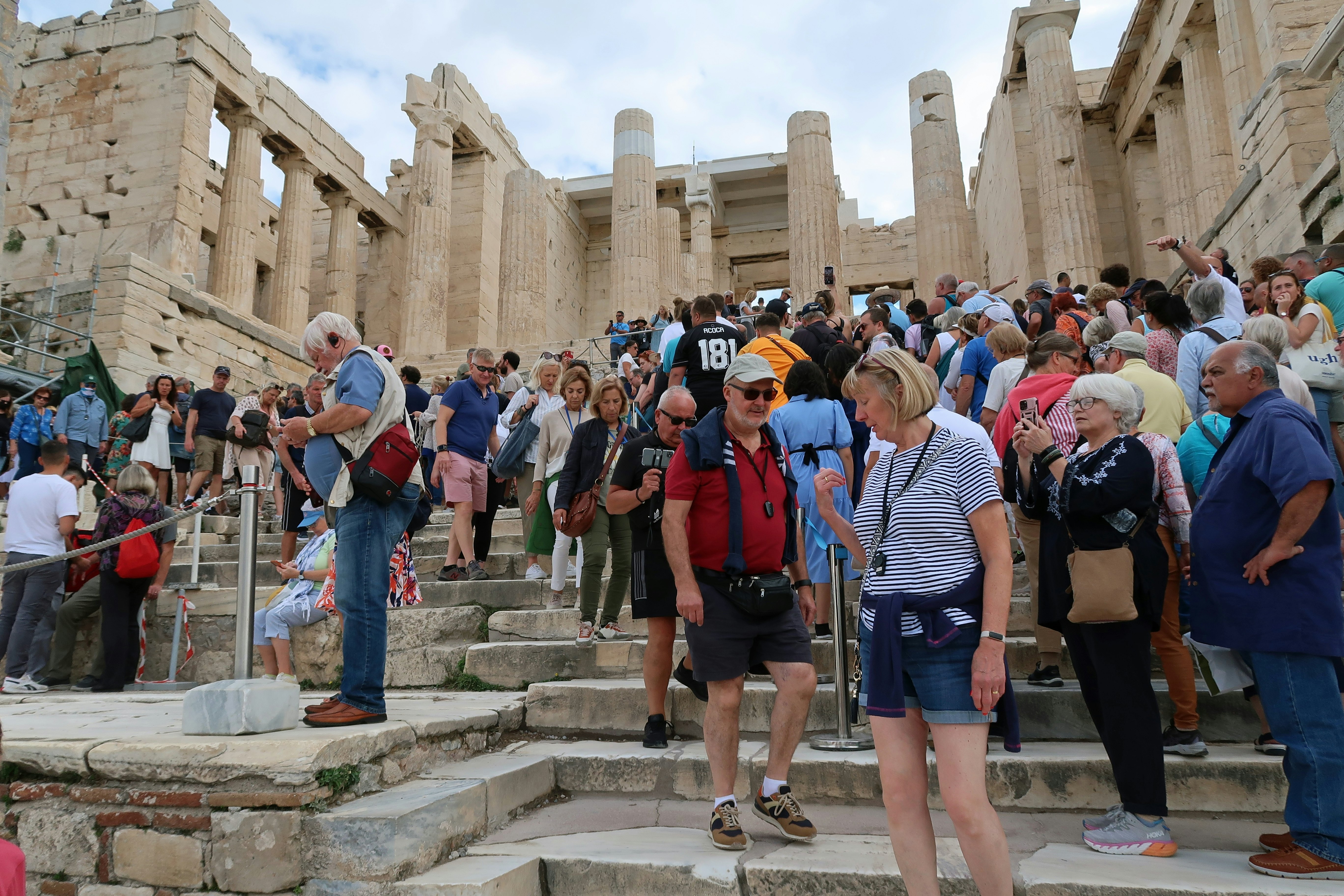 A large number of people walk on steps through an ancient site on a hot day. Columns and walls are visible in the background.