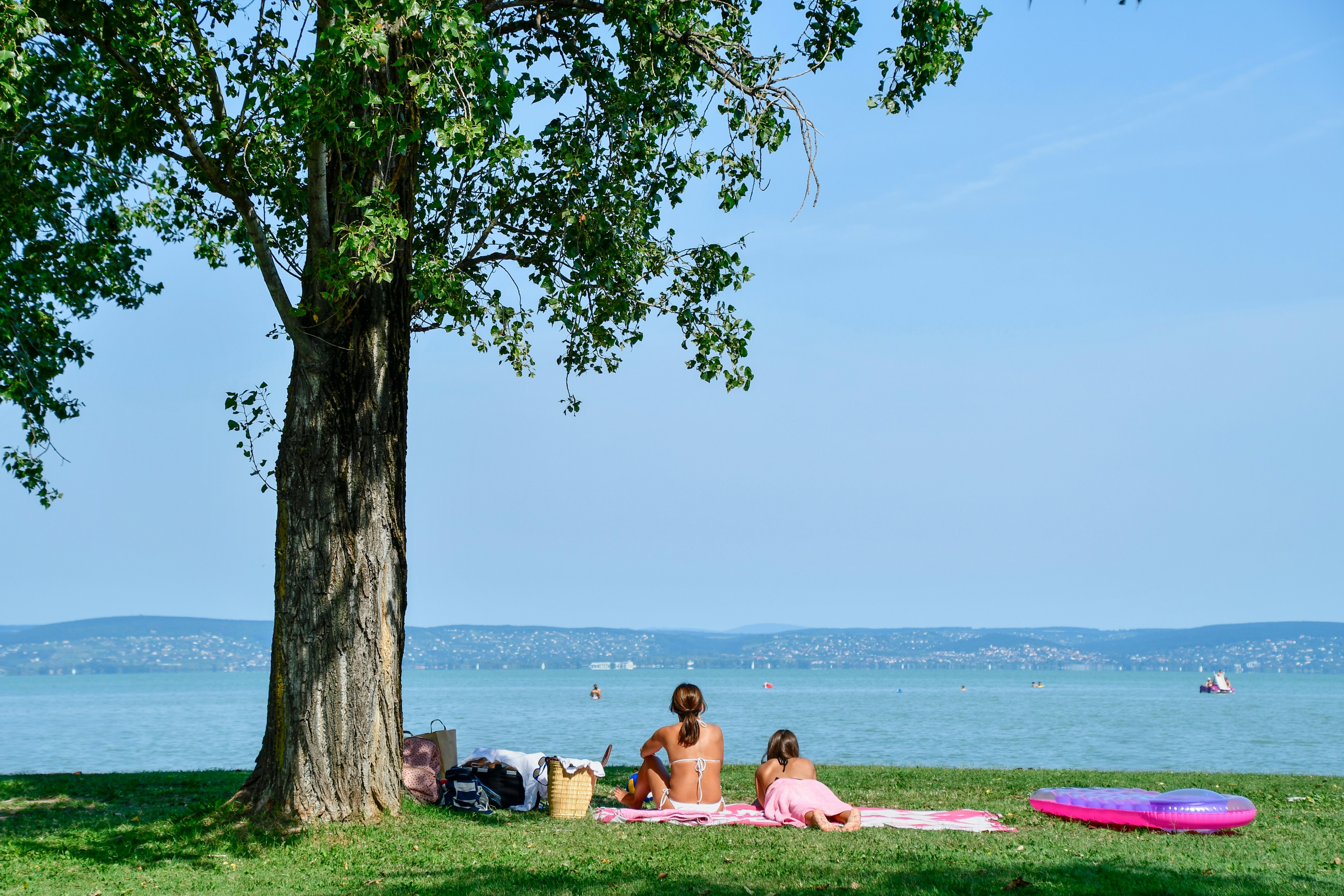 Two women in bathing suits on a picnic blanket on green grass under a tree with a large lake in the background.