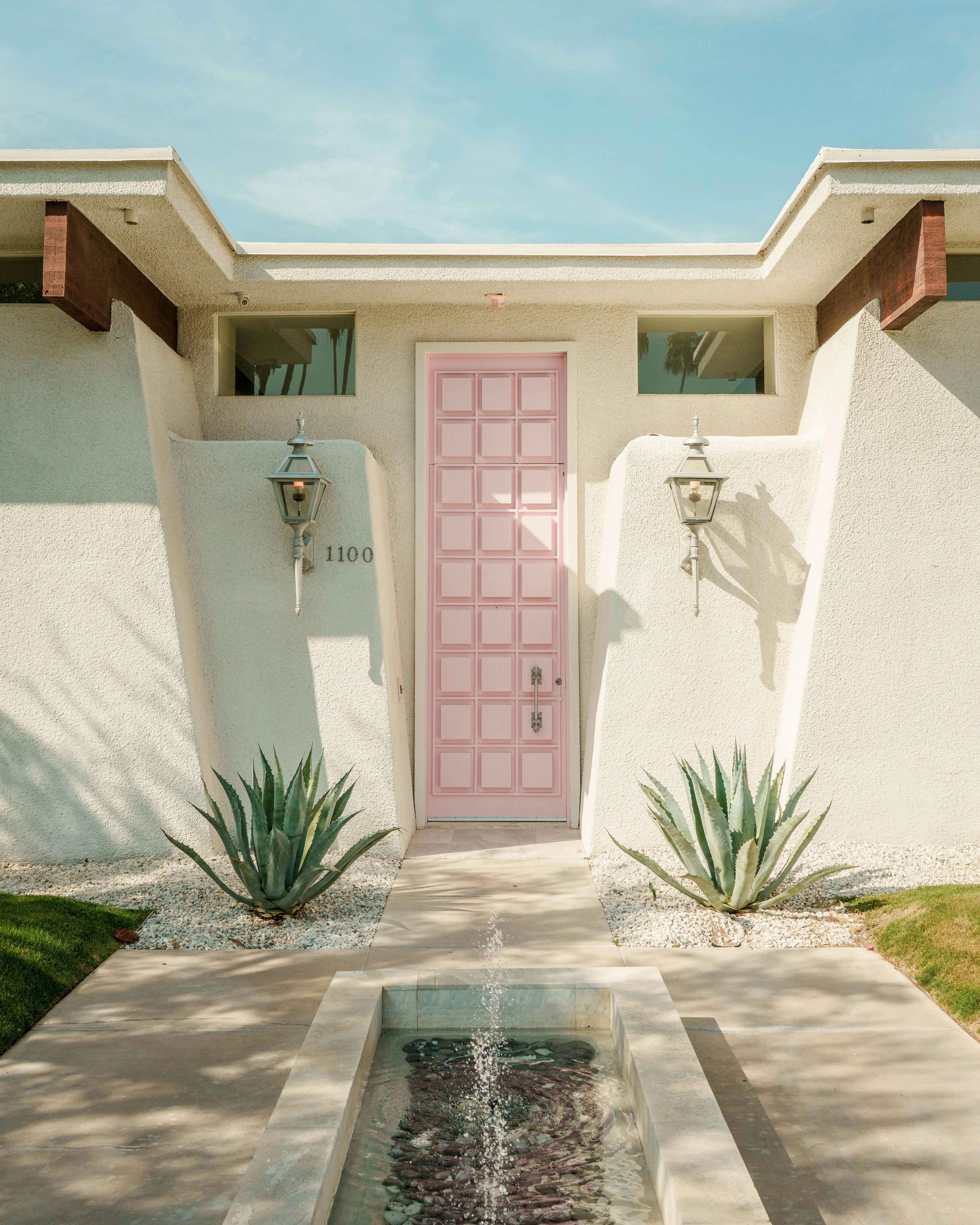 Close-up of a mid-century modern house with a pink door, plants, and a fountain.
