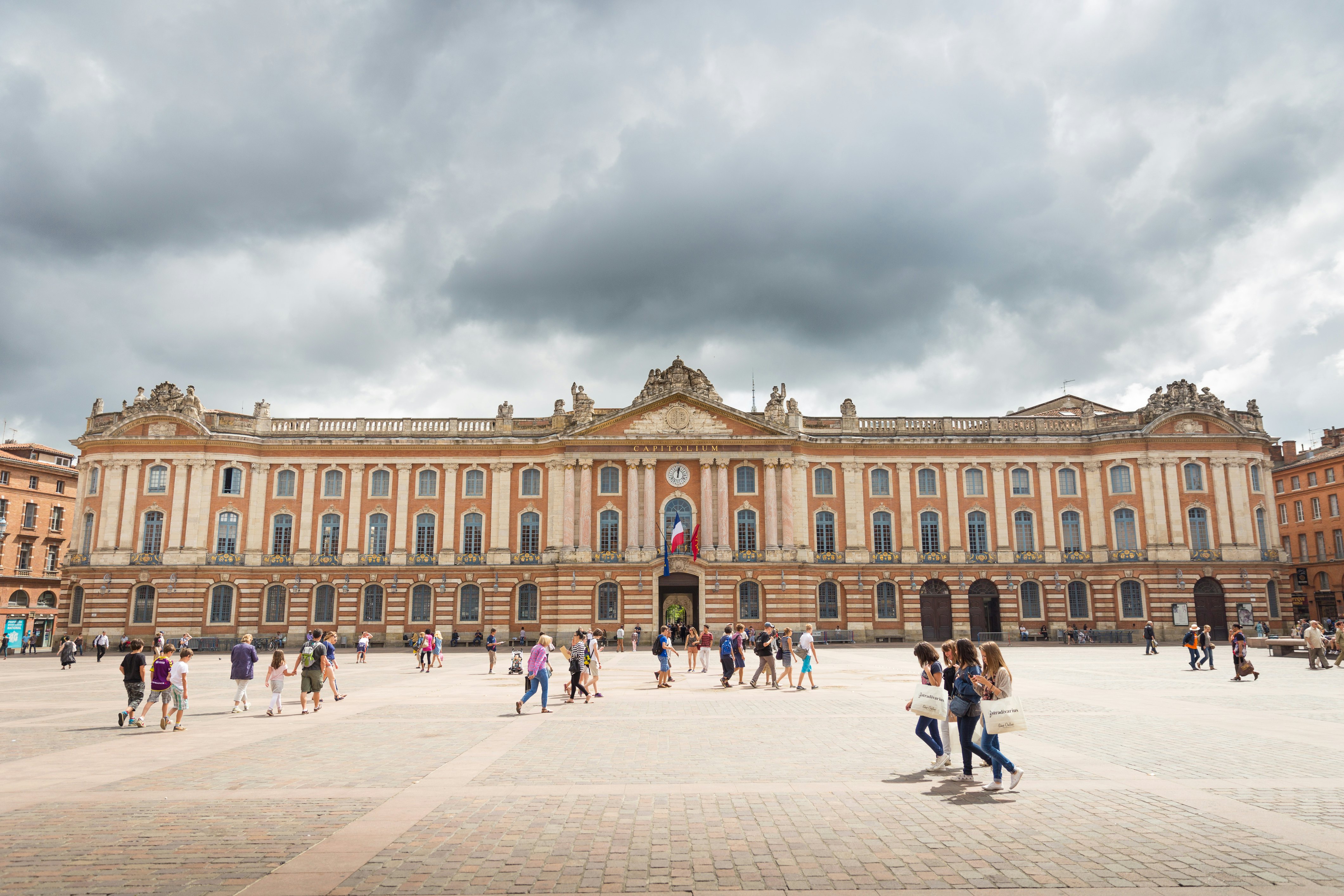People walk on a large square in front of a grand building made of pink and orange stone.