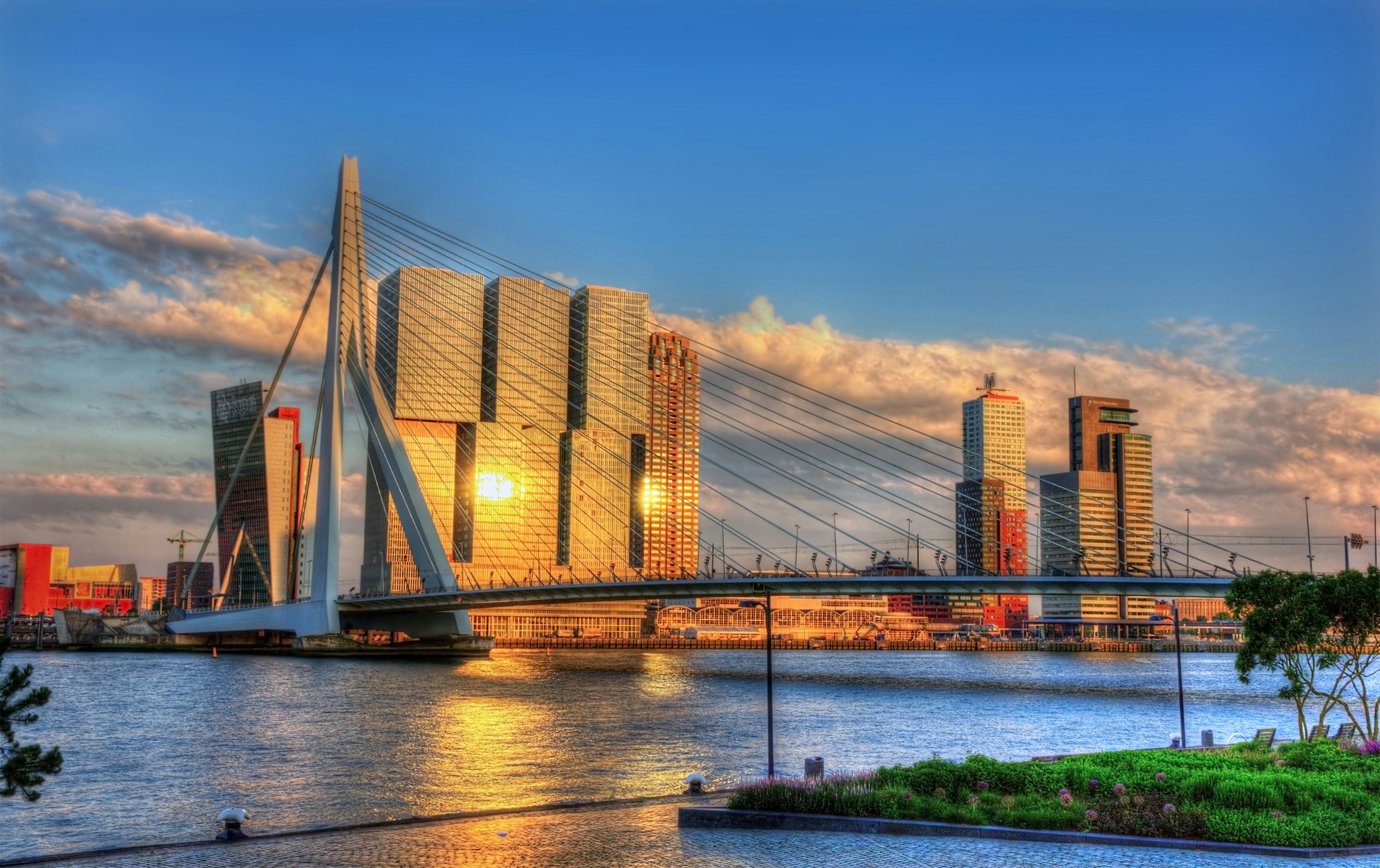 A suspension bridge over a river next to a large building of steel and glass reflecting the setting sun