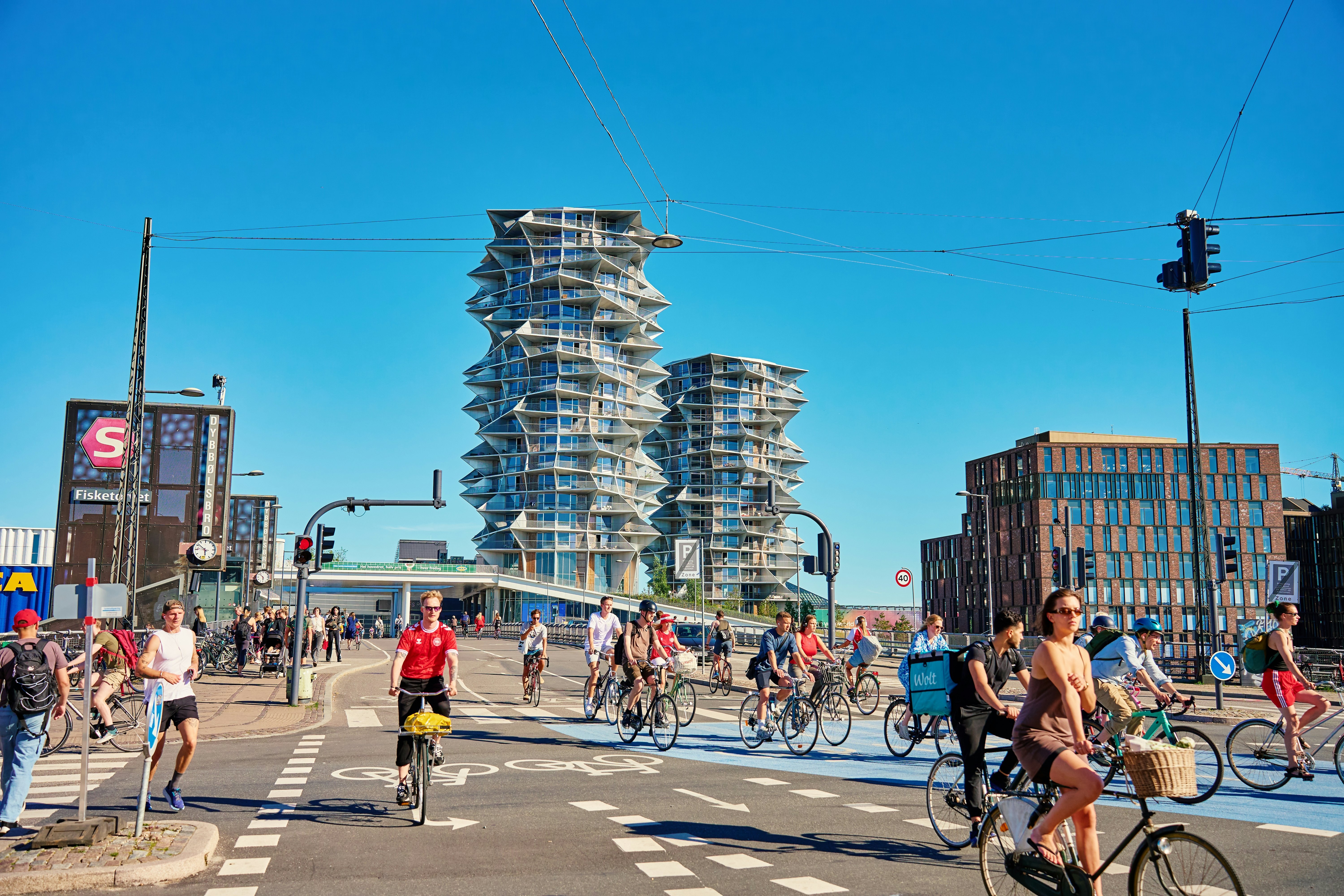 People riding bicycles on a sunny day in a street filled with bike lanes and modern buildings in the distance.