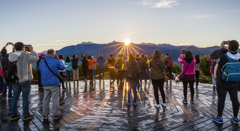 A group of tourists point cameras toward the sun rising over a line of mountain peaks.